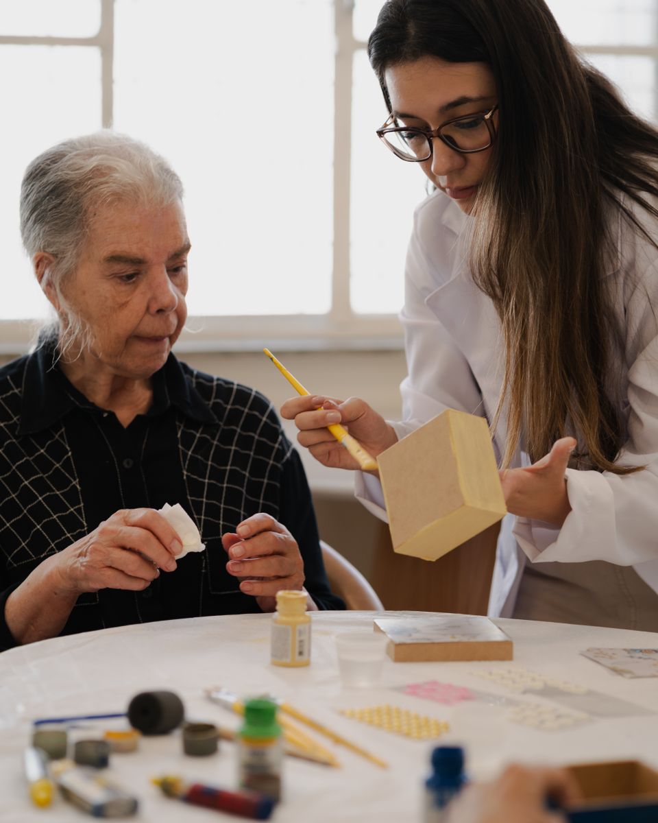 A caregiver helps an older woman with a craft project at a kitchen table.