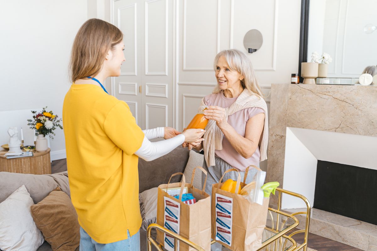 A caregiver hands a drink to an older woman in a living room.
